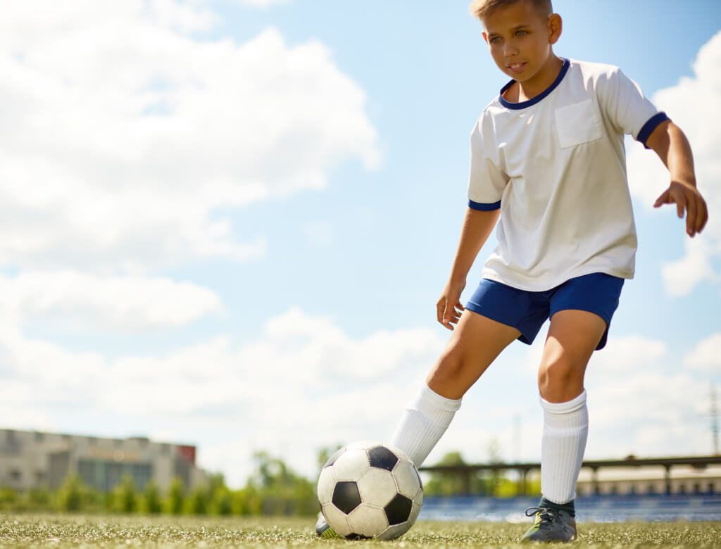 Ein kleiner Junge in einem weißen Hemd und blauen Shorts kickt energisch einen Fußball auf dem Spielfeld.