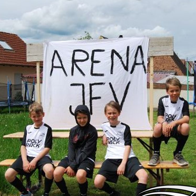 Eine Gruppe junger Jungen sitzt auf einer Bank, während ein Banner mit der Aufschrift "Arena JFV" im Hintergrund hängt.